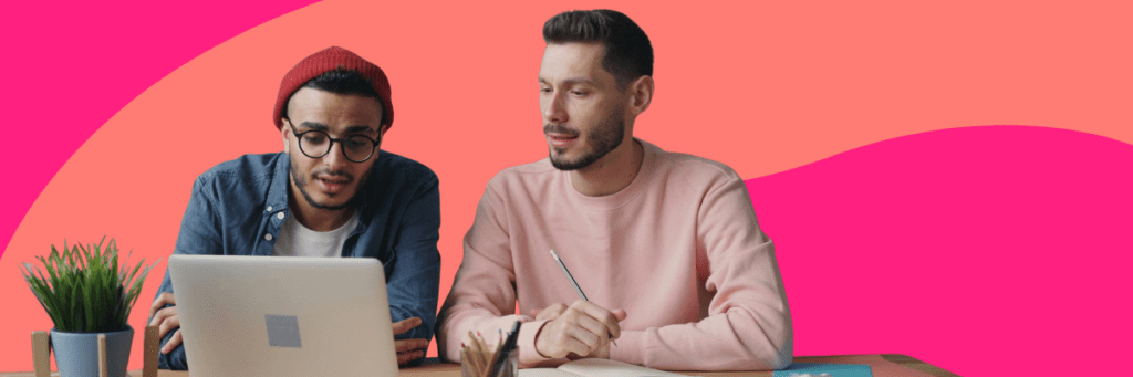 Two men sit at a desk looking at a laptop, with notebooks and a plant nearby, against a bright pink and coral abstract background.
