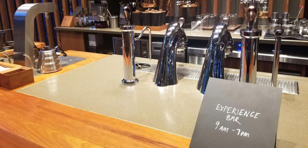 Coffee bar with silver brewing equipment on a wooden counter. A chalkboard reads "Experience Bar 9 AM - 7 PM.