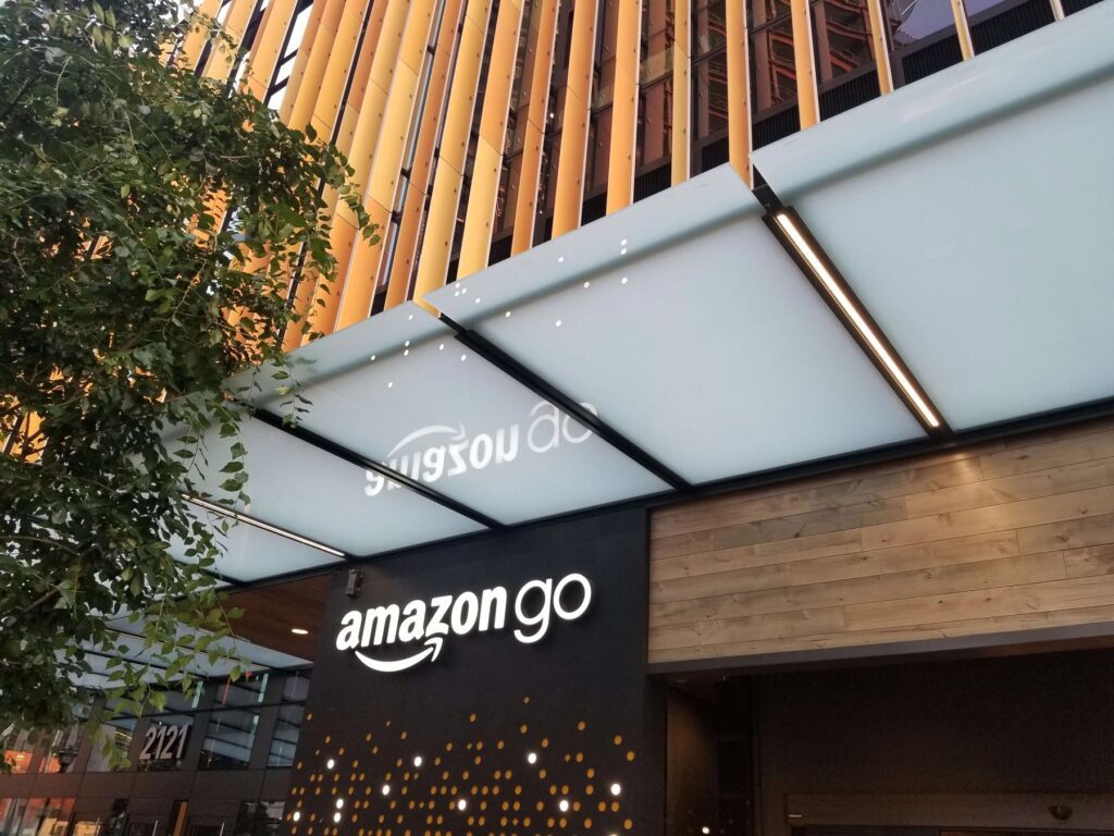 Amazon Go store entrance with a black sign displaying the logo. A modern overhang with lighting is above, and a building facade with vertical elements is in the background.