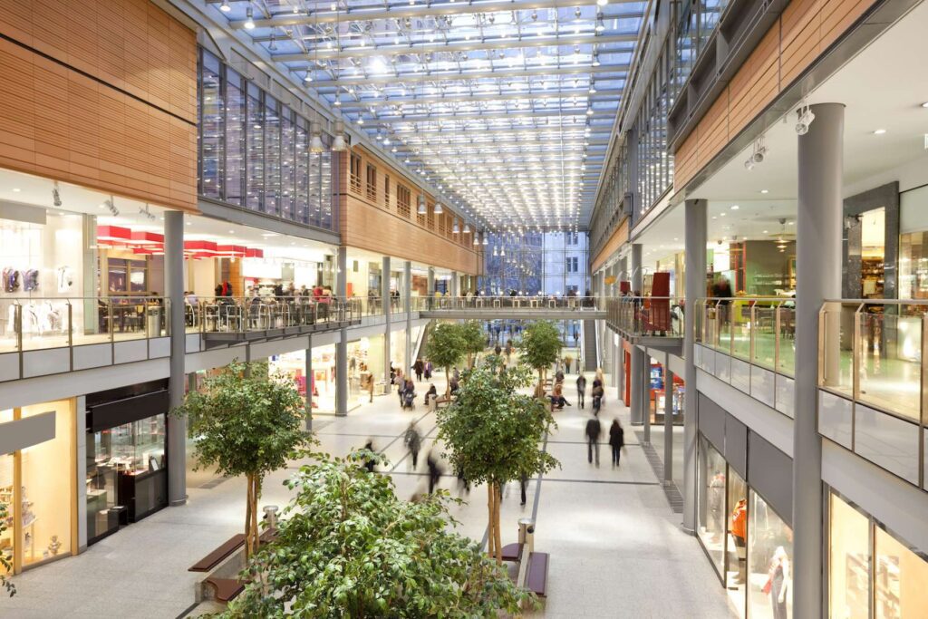 Interior of a modern shopping mall with two levels, glass roof, and indoor trees. People are walking and shopping, with various stores lining both sides.