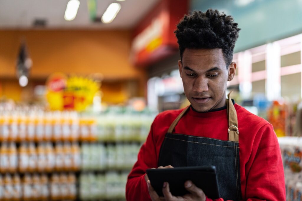 Store employee wearing an apron stands in front of shelves, looking at a tablet.