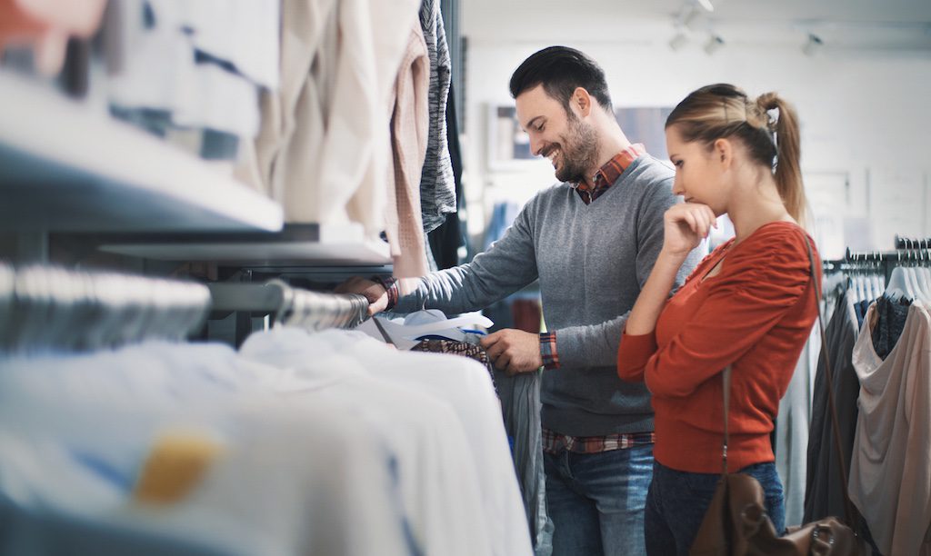 A man and a woman look at clothes on racks in a store; the man holds a shirt while the woman ponders.