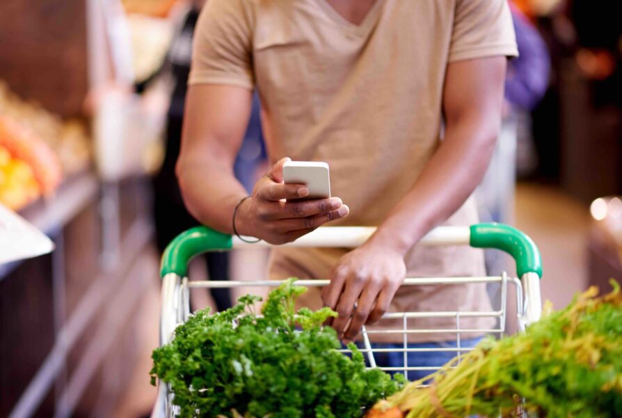 A person pushes a grocery cart filled with leafy greens while looking at a smartphone in a supermarket.