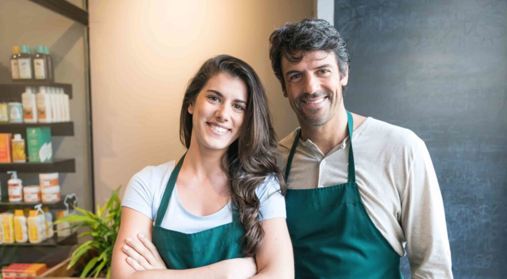 Two people wearing green aprons stand side by side in a store, smiling at the camera. Shelves with products and a chalkboard are visible in the background.