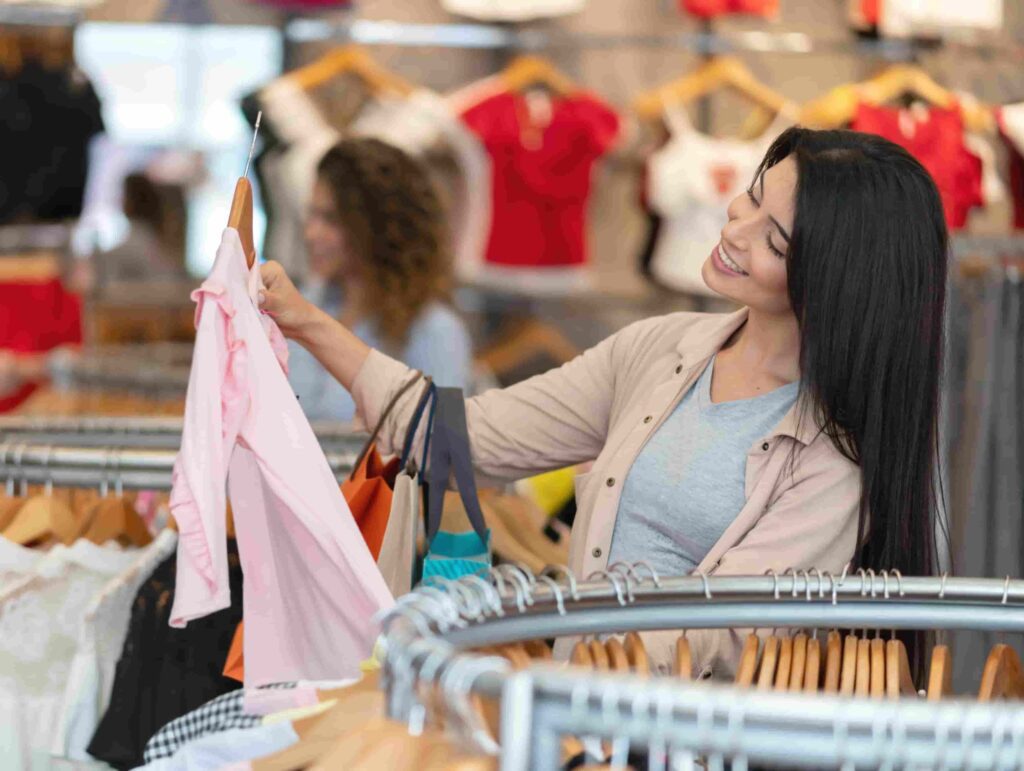 A woman stands by a clothing rack in a store, smiling as she holds up and examines a light pink shirt.