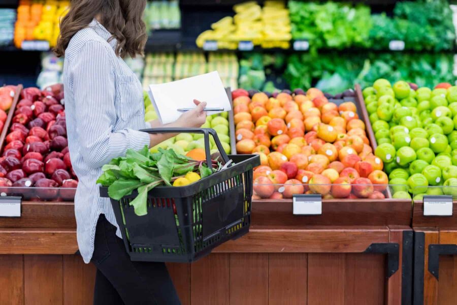 Person holding a shopping basket and notepad in front of a grocery store's fruit section with various apples and greens.