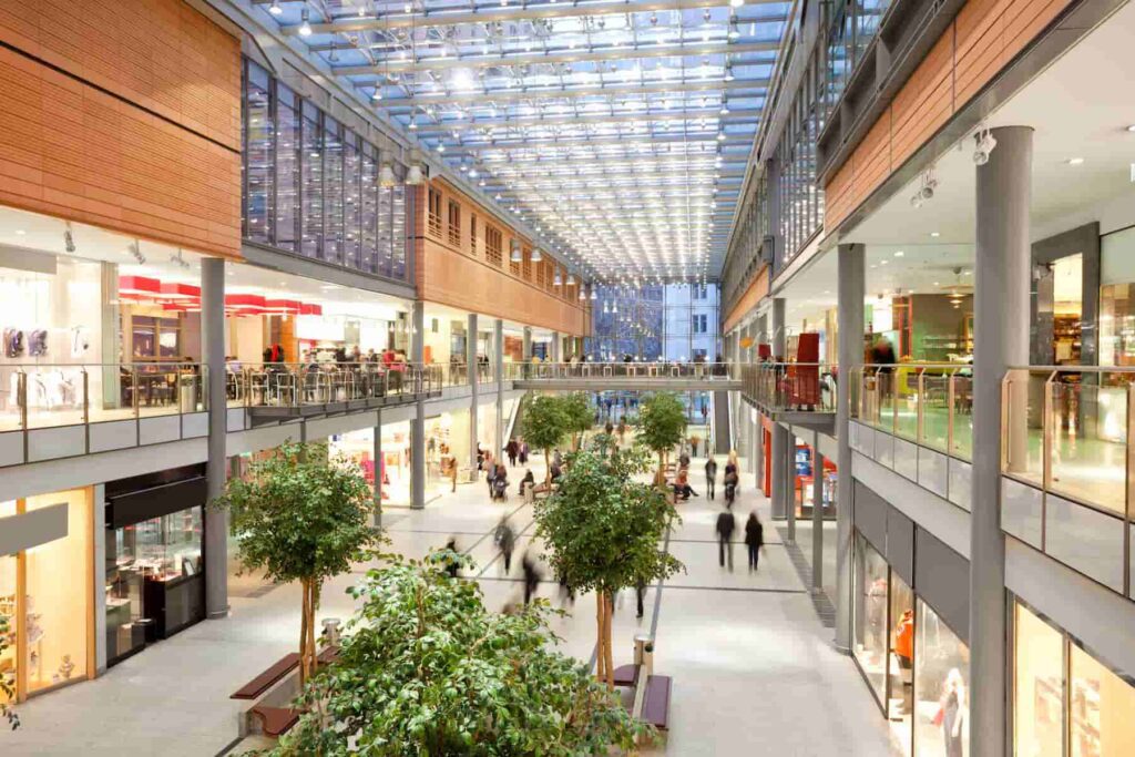Spacious indoor shopping mall with two floors, featuring shops, dining areas, and greenery under a glass ceiling. People are walking and browsing.