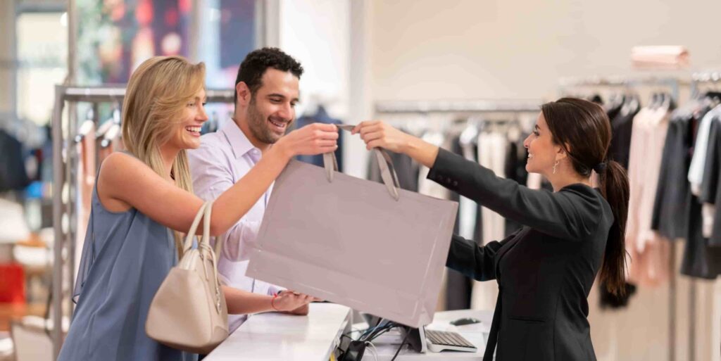 A woman accepts a shopping bag from a cashier while a man stands beside her at a clothing store counter.