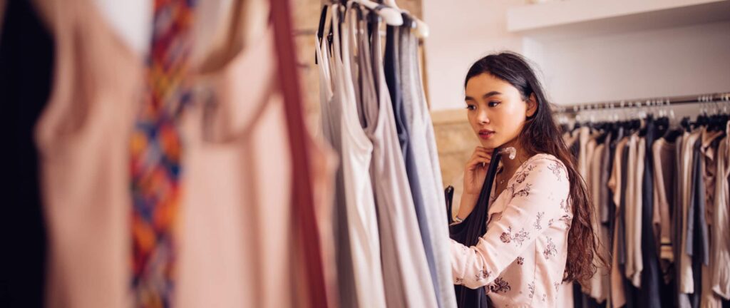 Woman browsing clothes on a rack in a store, wearing a light floral top, with a thoughtful expression.