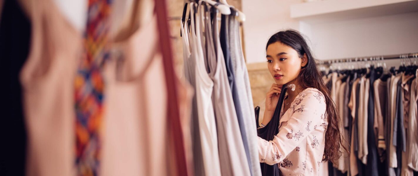Woman browsing clothes on a rack in a store, wearing a light floral top, with a thoughtful expression.