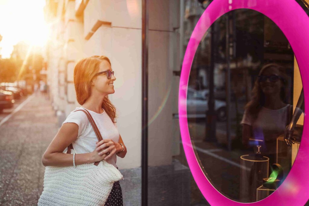 A woman wearing sunglasses and carrying a white bag looks into a store window with a pink neon oval frame at sunset.