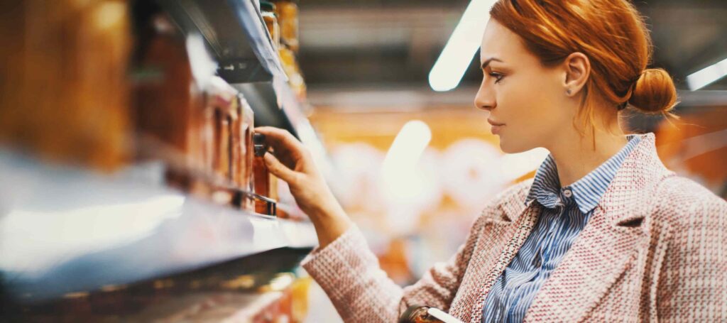 A woman with red hair wearing a coat and striped shirt selects an item from a grocery store shelf.