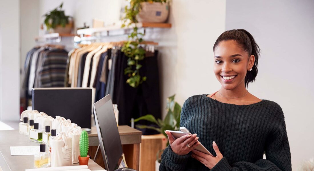 A woman stands behind a retail counter holding a tablet, smiling at the camera. Shelves with clothes and plants are visible in the background.