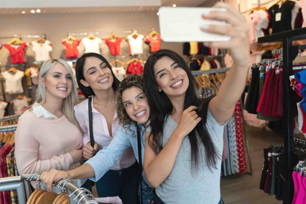 Four women standing together in a clothing store, smiling and taking a selfie with a smartphone. Clothing racks and shelves are visible in the background.
