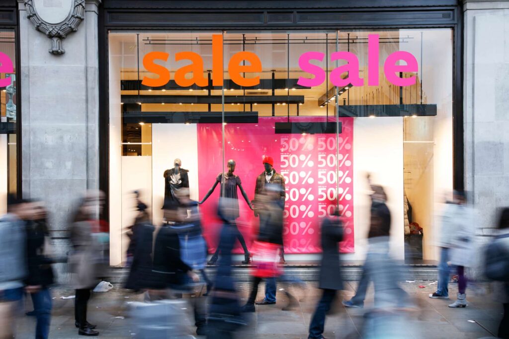 Busy street with people walking past a store displaying "sale" signs and mannequins. Large red posters show "50% off" inside the window.