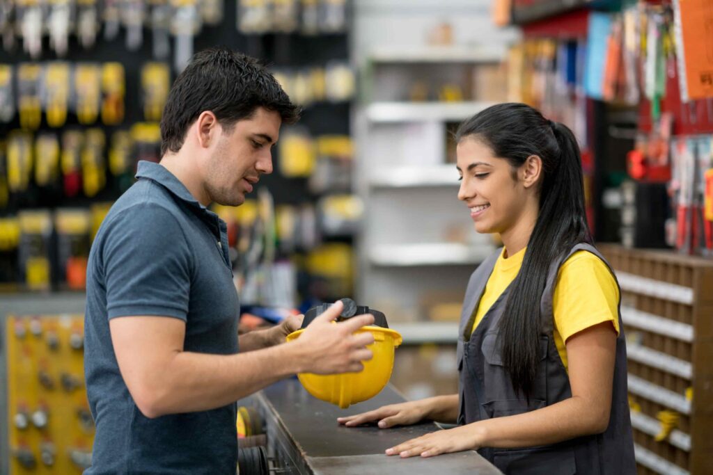 A man and a woman stand at a hardware store counter. The man holds a yellow hard hat and gloves while talking to the woman, who is wearing a vest and smiling.