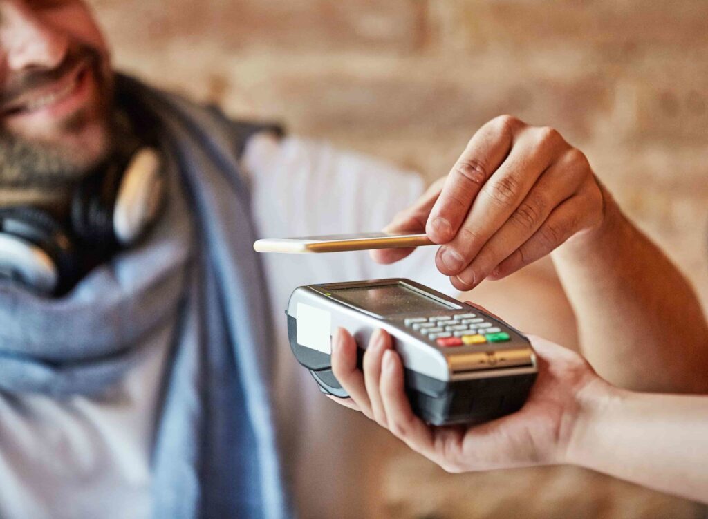 A person holds a smartphone over a card reader to make a contactless payment while another person holds the payment terminal.