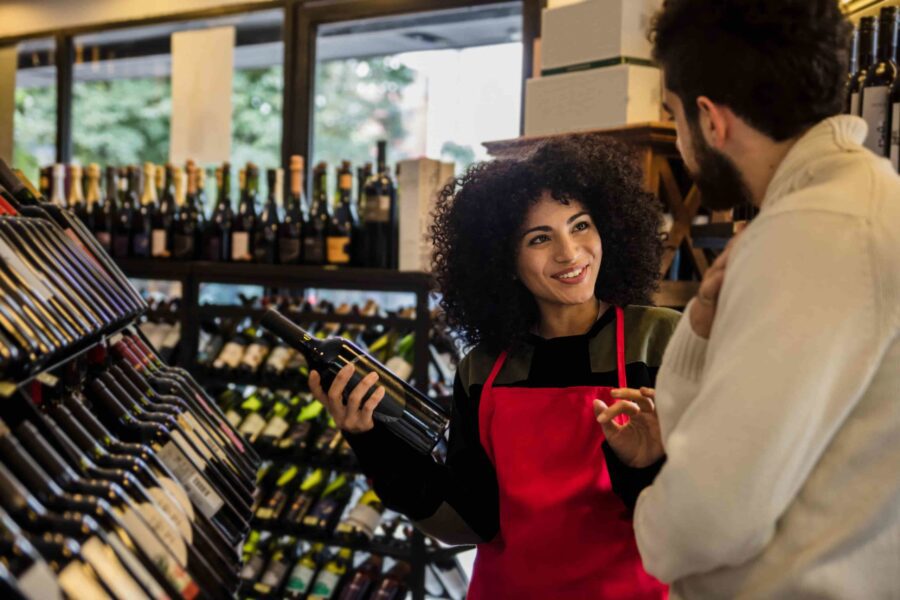 A store employee in a red apron holds a wine bottle and talks to a customer in a wine shop with shelves full of bottles.