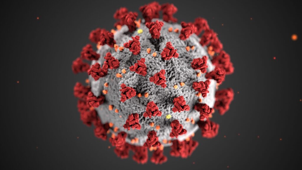 3D illustration of a coronavirus particle, showing a spherical gray structure with red spike proteins and small orange dots on a dark background.