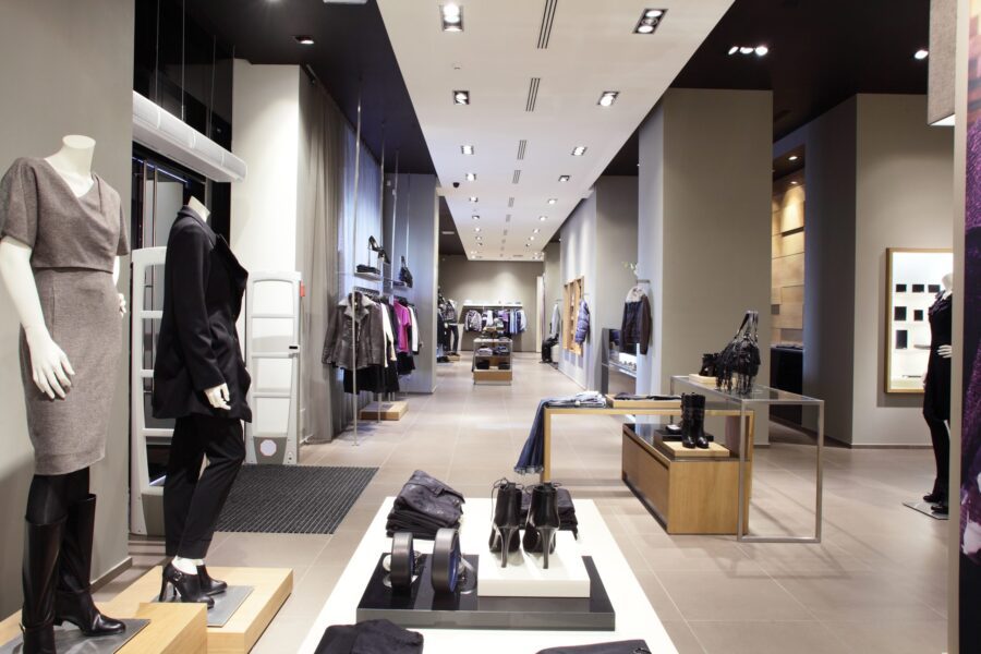 Modern clothing store interior with mannequins dressed in casual and business attire, display tables with folded clothes, and racks of garments lining the walls.