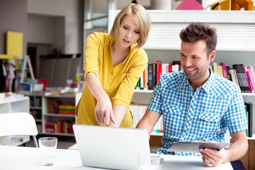A woman standing and pointing at a laptop screen while a man sits beside her holding a tablet; both are in a modern office setting.