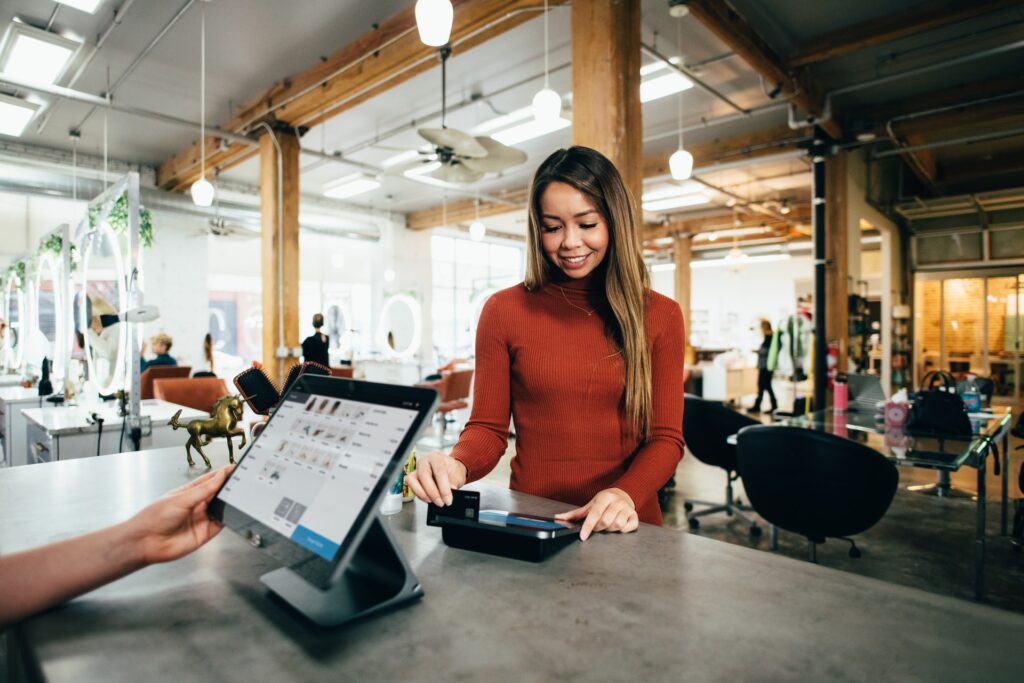 A woman in a red sweater completes a transaction using a payment card on a card reader in a modern, well-lit store. A cashier is on the other side of the counter using a touchscreen device.