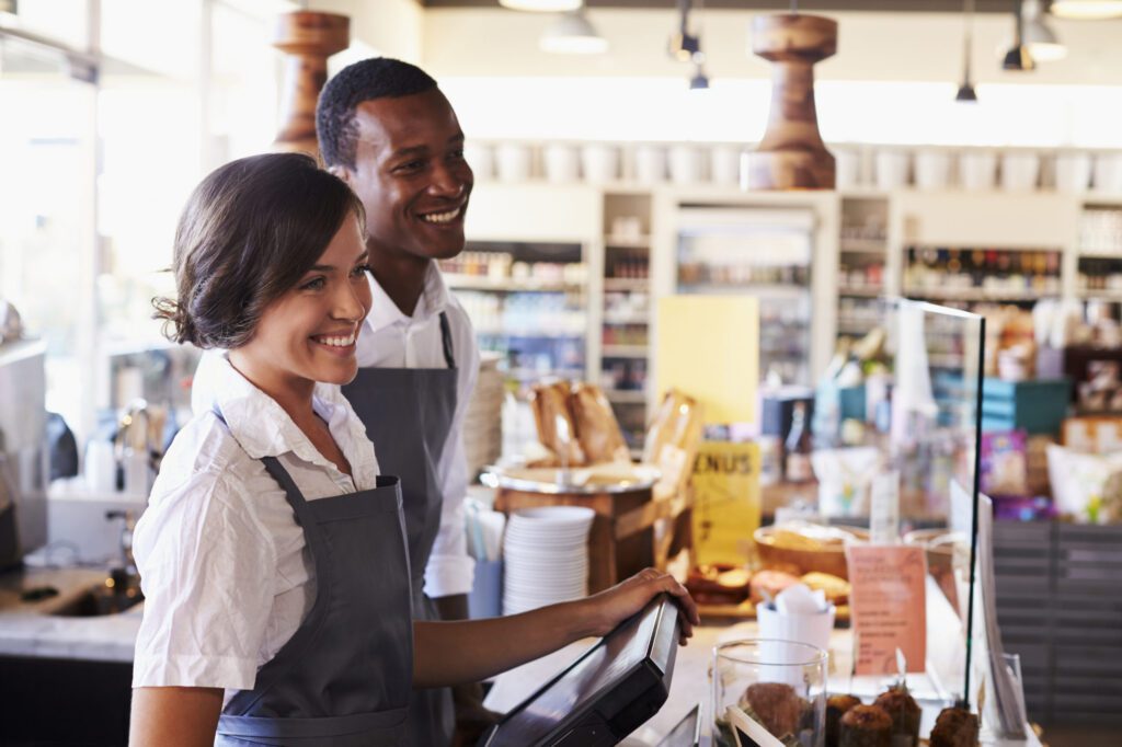 Two employees in aprons stand behind a counter at a café or bakery, smiling while operating the cash register. Shelves and baked goods are visible in the background.