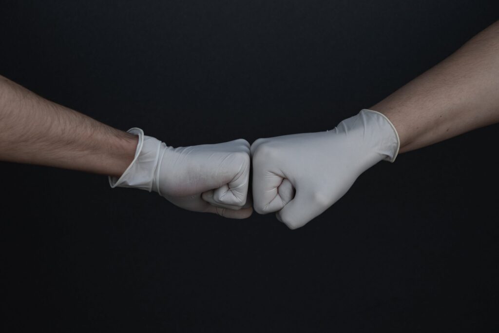 Two people wearing white disposable gloves bump fists against a black background.