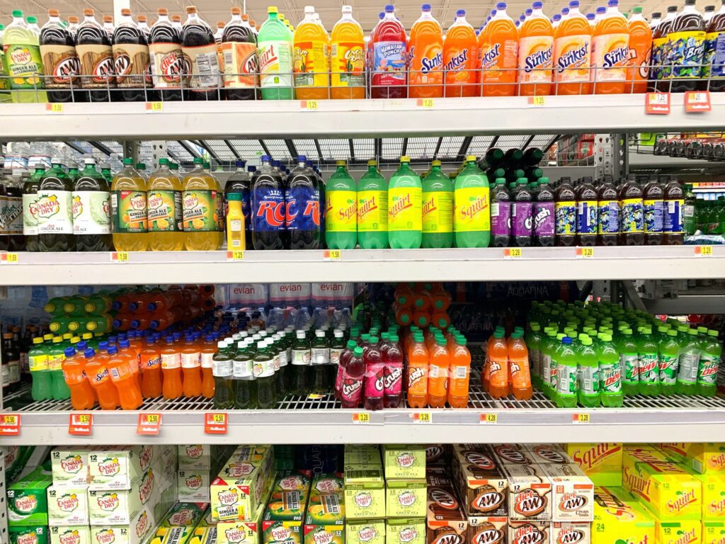 Shelves in a store stocked with various brands and flavors of sodas in plastic bottles, arranged in rows by color and type.
