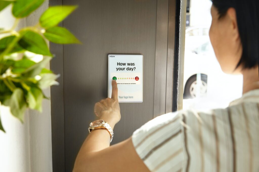 A person presses a green button on a wall-mounted feedback device displaying "How was your day?" with a satisfaction scale.