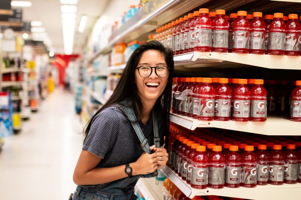A person wearing glasses and a striped shirt smiles while standing next to shelves stocked with bottles of red Gatorade in a grocery store.