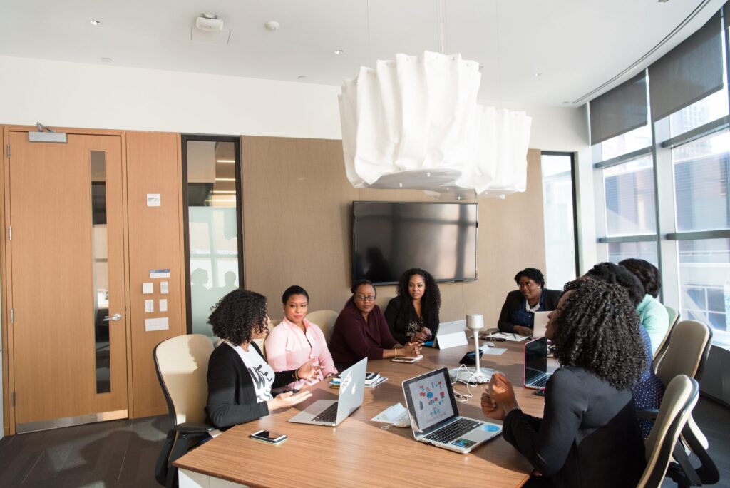 A group of people sit around a conference table with laptops and notepads, engaged in a meeting in a modern office room.