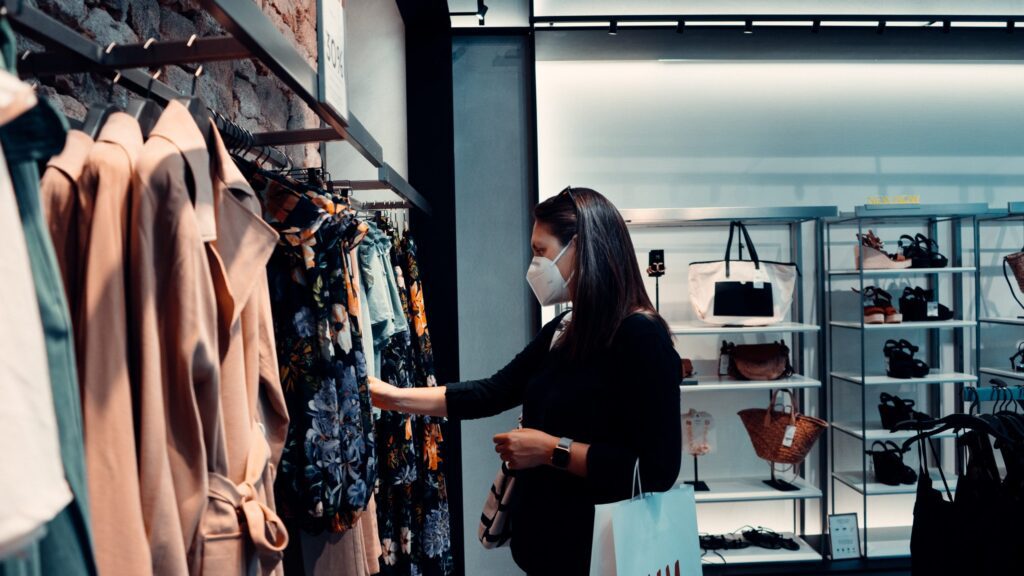 A woman wearing a face mask shops for clothes in a store, browsing through a rack of garments with shopping bags in hand.