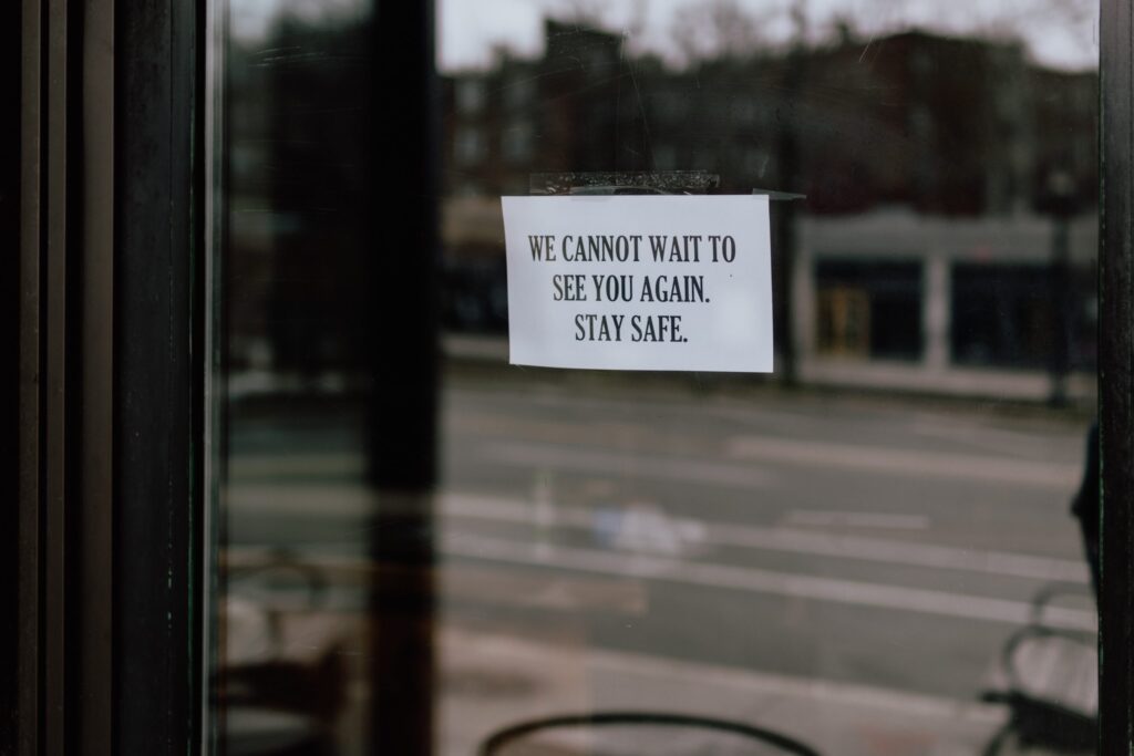A paper sign taped to a window reads, "WE CANNOT WAIT TO SEE YOU AGAIN. STAY SAFE." An empty street and blurred buildings are visible outside.