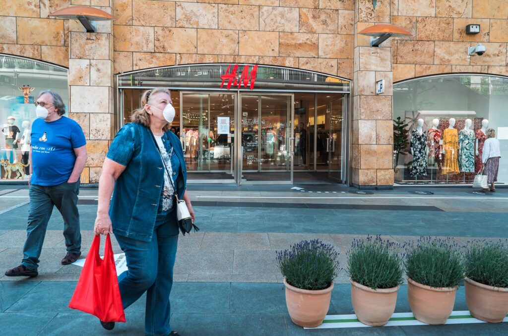 Two people wearing face masks walk past an H&M store entrance on a city street; mannequins in dresses are visible inside the store.