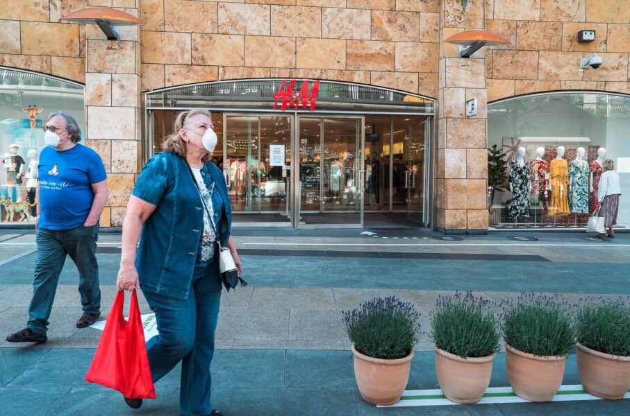 Two people wearing face masks walk past an H&M store entrance on a city street; mannequins in dresses are visible inside the store.