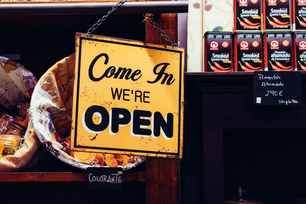 A yellow "Come In We're OPEN" sign hangs in a store, with jars on shelves and a price tag for smoked paprika visible nearby.