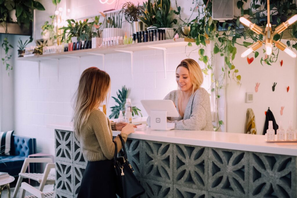 A woman stands at a modern salon reception desk, speaking with a receptionist who is using a point-of-sale system. Shelves with products and plants decorate the background.