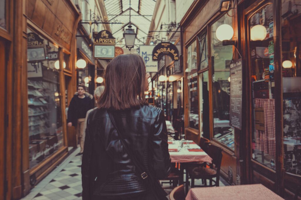 A person in a black leather jacket walks through a narrow, indoor shopping arcade lined with shops and lit by round wall lamps.