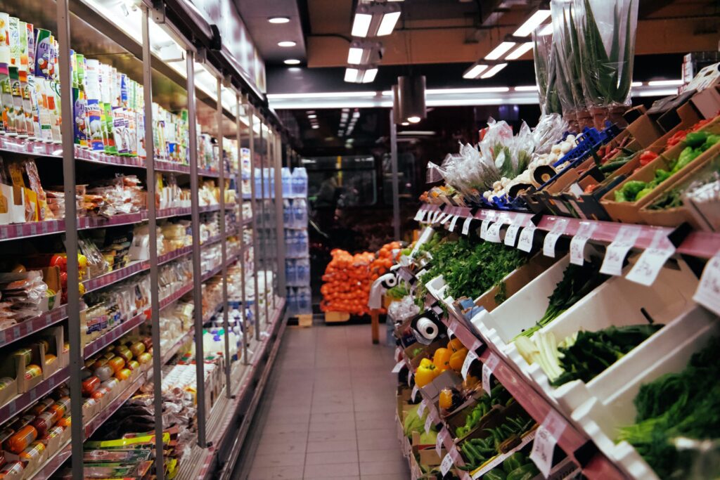 Grocery store aisle with refrigerated shelves on the left holding packaged foods and a produce section on the right displaying various fresh vegetables.