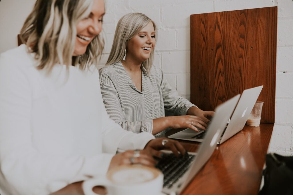 Two women sit at a wooden counter, each working on a laptop. One has a coffee cup in front of her. Both are smiling and appear engaged in their tasks.