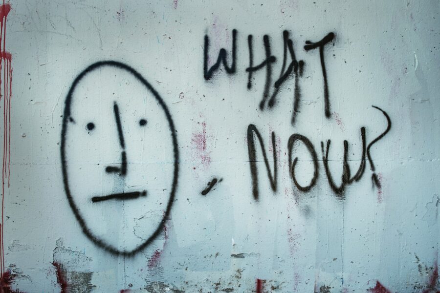 Graffiti on a wall depicts a simple face with two dots for eyes and a line for a mouth, next to the words "WHAT NOW" in black spray paint.