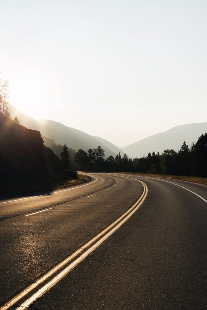 A paved road with double yellow lines curves through a mountainous landscape under a hazy sky, with sunlight illuminating the scene from the left.
