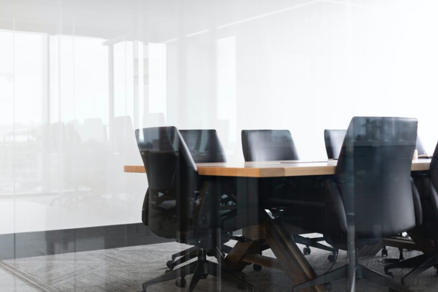 A modern, empty conference room with a wooden table and black chairs, seen through a glass wall. Natural light enters from large windows in the background.