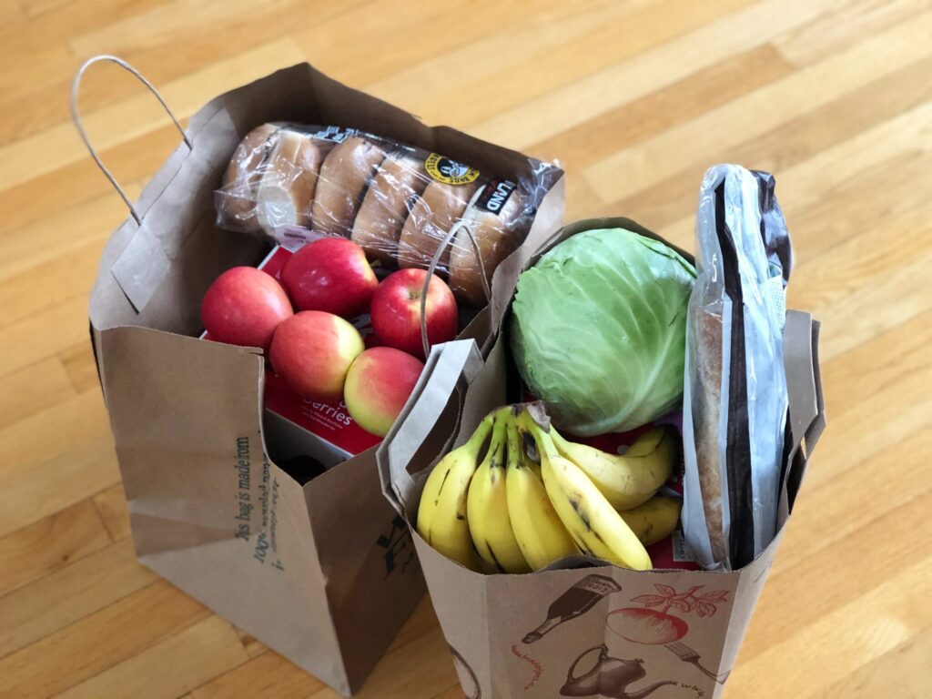 Two paper grocery bags on a wooden floor, filled with apples, bananas, cabbage, a loaf of bread, a pack of bagels, and other assorted groceries.