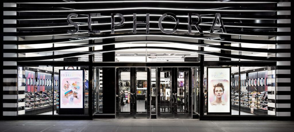 A Sephora store entrance with large glass doors flanked by display windows showcasing beauty products and promotional posters.