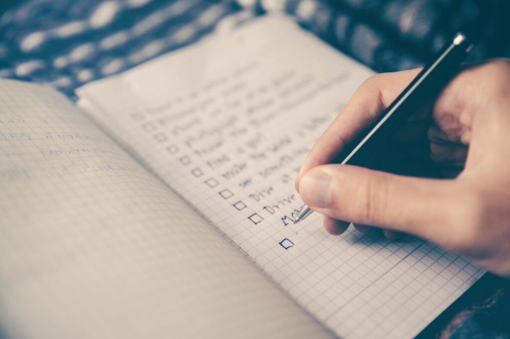 A person writes on a checkered notebook, filling out a checklist with several completed and some empty boxes.