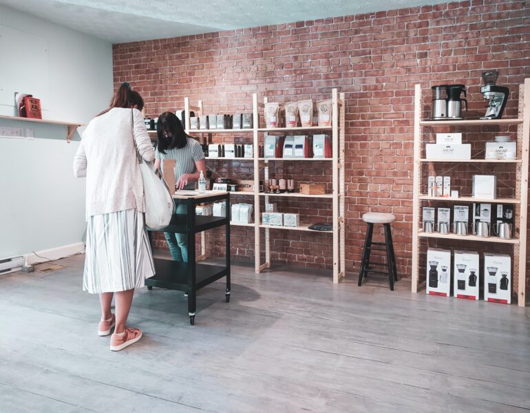 Two people stand at a table in a store with shelves displaying various coffee products and items against a brick wall.