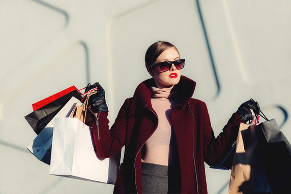 A person in a burgundy coat and sunglasses holds multiple shopping bags, standing against a light-colored, geometric background.