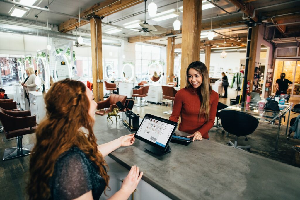 Two women interacting at a modern salon reception desk. One is behind the counter with a tablet; the other is facing her, gesturing towards the screen. The salon is spacious and well-lit.